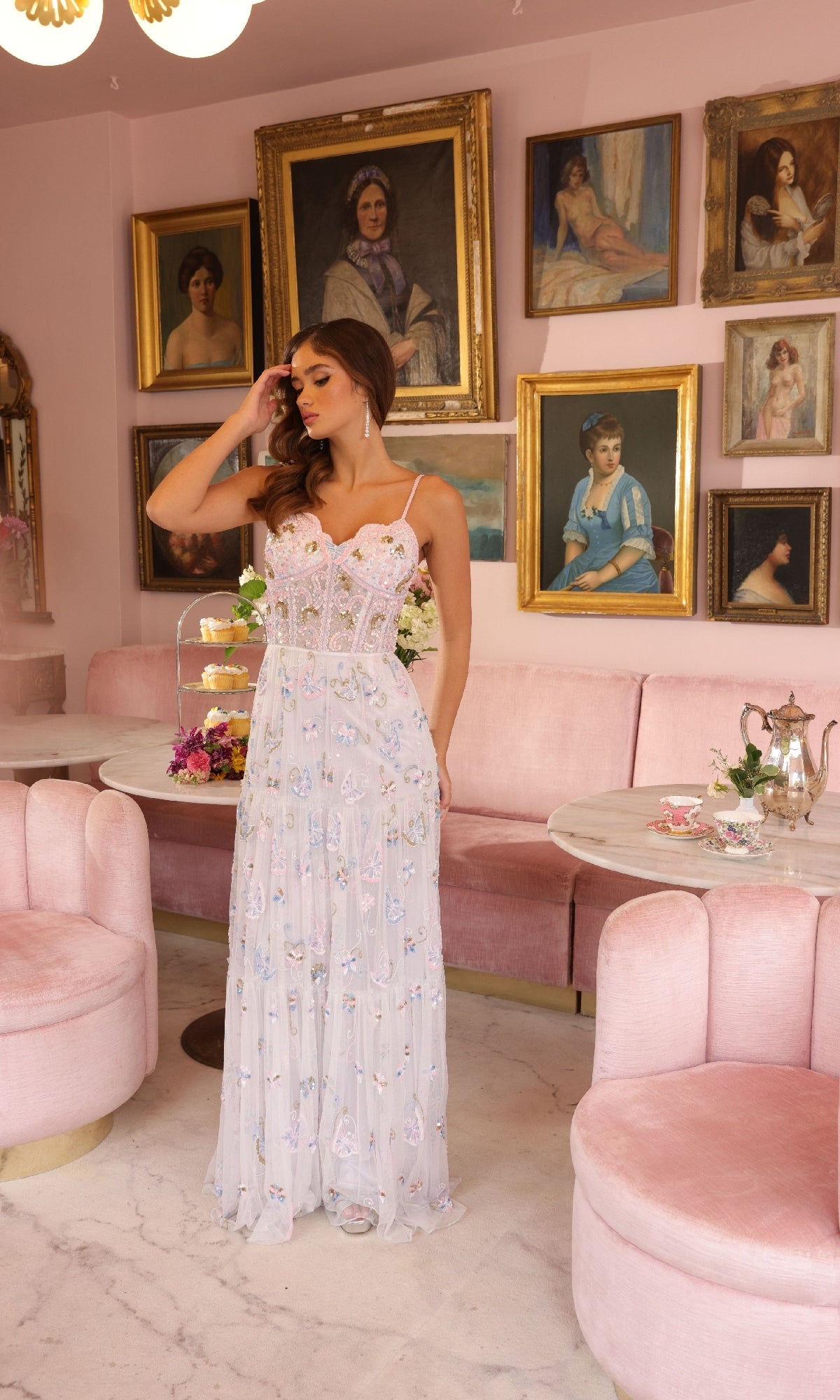 A woman in an Ava Presley 47044 long prom dress stands in a pink room with round seating, marble tables, and framed portraits on the wall.